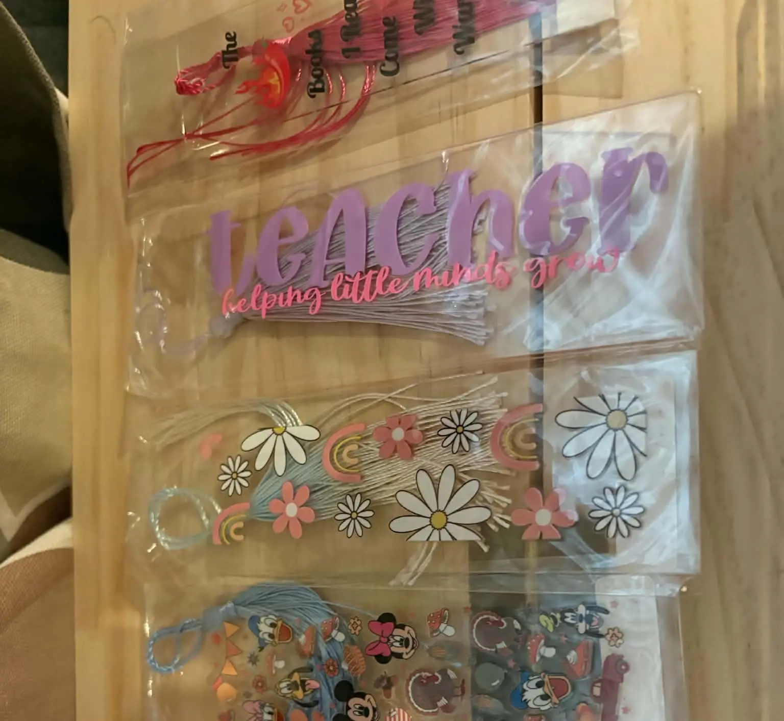A stack of clear tassel bookmarks, including a Teacher design and a daisy and rainbow design